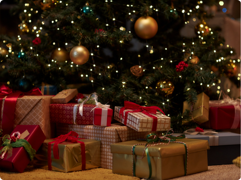 Festlich geschmückter Weihnachtsbaum mit warmen Lichtern und zahlreichen verpackten Geschenken in Rot-, Gold- und Grüntönen darunter.